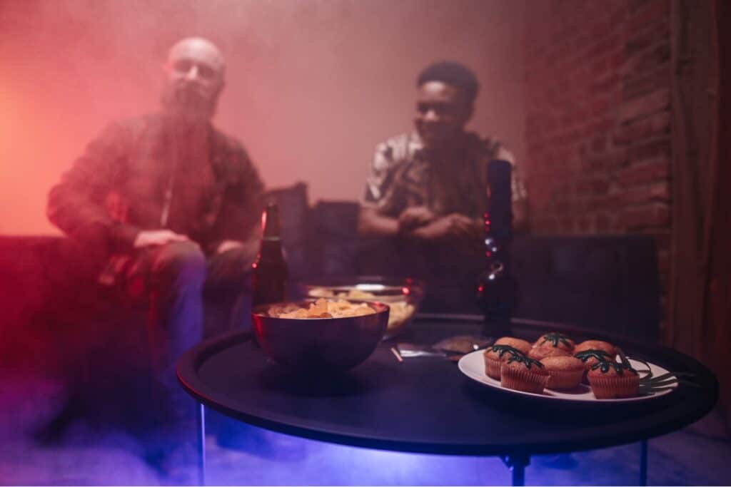 two individuals looking at marijuana munchies on a table in a room filled with smoke